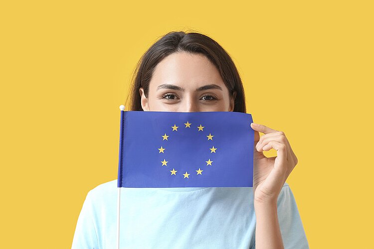 Junge Frau vor gelben Hintergrund, die eine Europafahne vor ihr Gesicht hält.